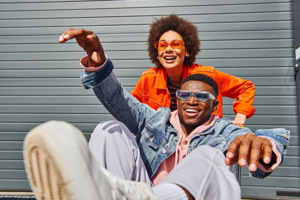 Positive afrikanisch-amerikanische Frau in Sonnenbrille und heller Kleidung hat Spaß mit stilvollen besten Freund in Jeansjacke sitzt im Einkaufswagen auf der städtischen Straße, Freunde mit stilvollem Ambiente — Stockfoto