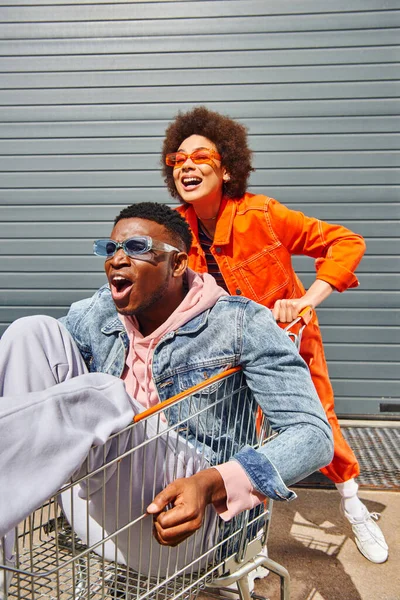 Sorridente giovane donna afroamericana in abito luminoso e occhiali da sole divertirsi con elegante e spaventato migliore amico seduto nel carrello della spesa vicino edificio sulla strada urbana, amici con atmosfera elegante — Foto stock