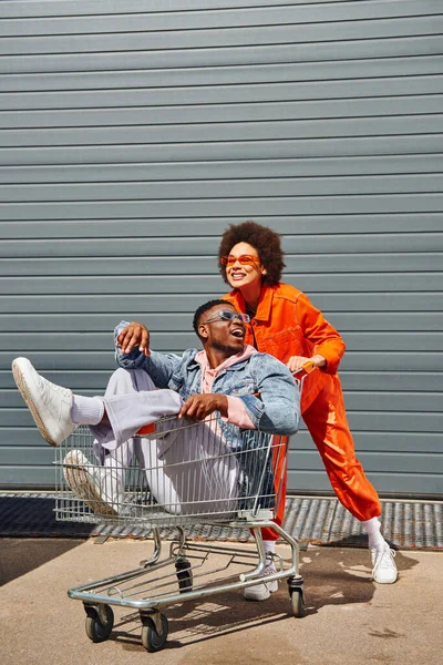 Young and excited african american best friends in sunglasses and bright outfits having fun with shopping cart and spending time near building on urban street, friends with stylish vibe — Stock Photo