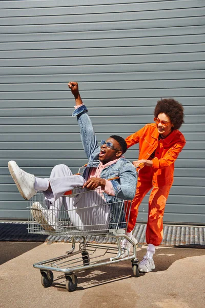 Full length of smiling young african american woman in sunglasses and bright outfit having fun with excited best friend sitting in shopping cart near building on urban street, stylish friends in city — Stock Photo