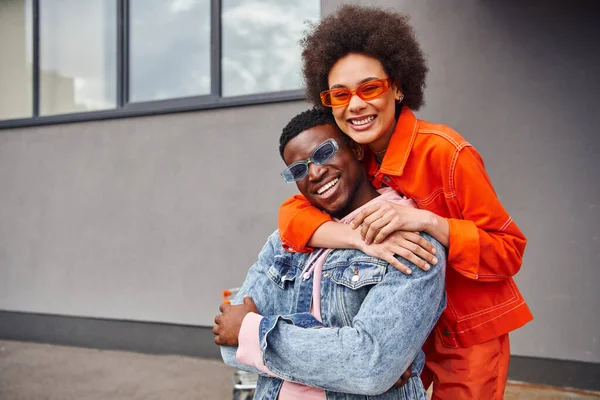 Smiling young african american woman in sunglasses and modern outfit embracing best friend and looking at camera while standing near building on urban street, stylish friends in city — Stock Photo