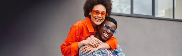 Positiva jovem afro-americana em óculos de sol e roupa brilhante abraçando melhor amigo e olhando para a câmera enquanto está perto de edifício na rua urbana, amigos elegantes na cidade, banner — Fotografia de Stock