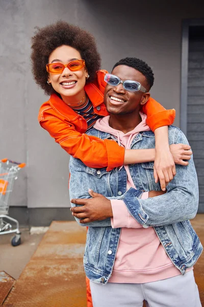 Positive young and trendy african american woman in sunglasses and bright outfit embracing best friend in denim jacket and standing near building on urban street, stylish friends in city — Stock Photo