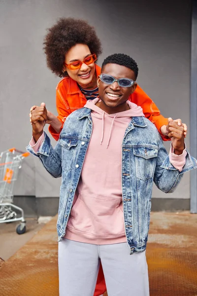 Happy young african american woman in sunglasses holding hands of trendy best friend in denim jacket while standing together near building on urban street, stylish friends in city concept — Stock Photo