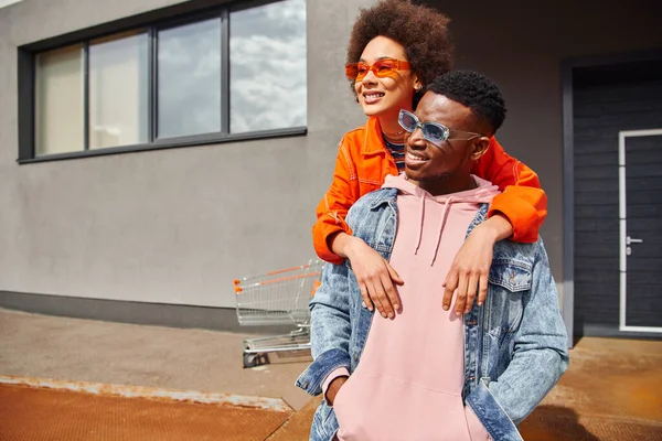 Lächelnde junge afrikanisch-amerikanische Frau in Sonnenbrille und heller Kleidung umarmt modische beste Freundin in Jeansjacke, während sie Zeit auf urbanen Straßen verbringt, stilvolle Freunde im Stadtkonzept — Stockfoto