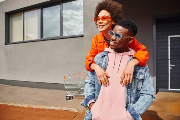 Sorrindo jovem afro-americana em óculos de sol e roupas brilhantes abraçando melhor amigo na moda em jaqueta jeans enquanto passa o tempo na rua urbana, amigos elegantes no conceito de cidade — Fotografia de Stock