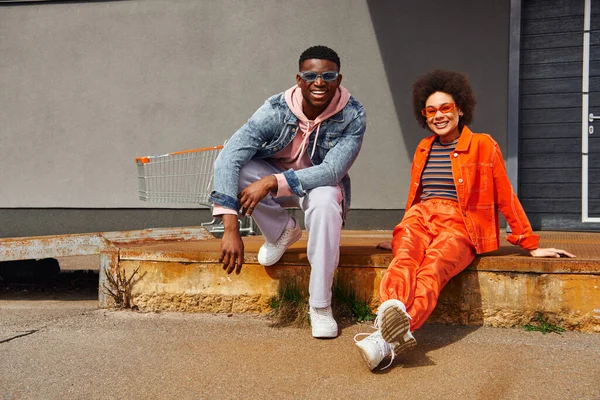 Full length of cheerful and trendy young african american best friends in sunglasses and modern outfits looking at camera while posing on rusty stairs near building outdoors, stylish friends in city — Stock Photo