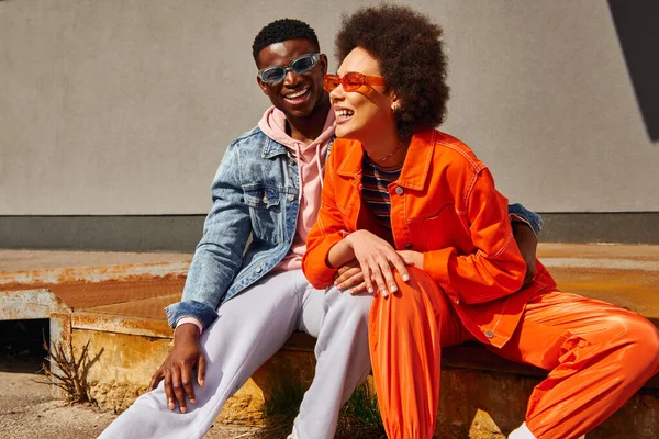 Fröhliche junge afrikanisch-amerikanische beste Freunde in trendigen Outfits und Sonnenbrille, die lachen und Spaß haben, während sie auf rostigen Treppen in der Nähe von Gebäuden an der Stadtstraße sitzen, trendige Freunde in urbanen Umgebungen — Stockfoto