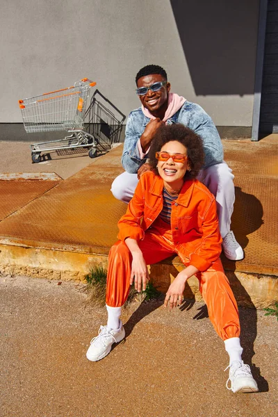 Comprimento total de alegre jovem afro-americana em óculos de sol e roupa brilhante sentado em escadas enferrujadas perto de elegante melhor amigo e edifício na rua urbana, amigos da moda em ambientes urbanos — Fotografia de Stock