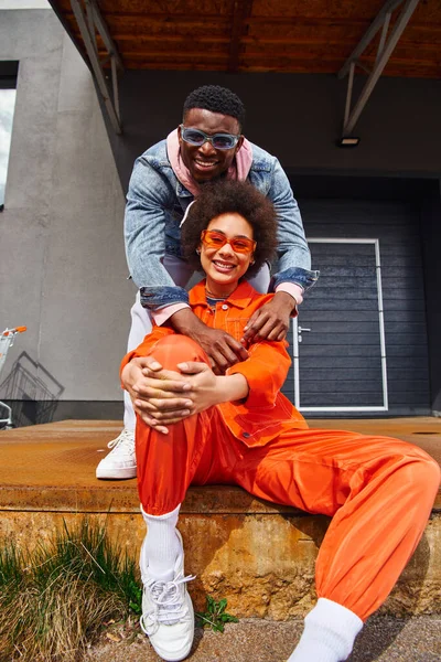 Homem americano africano jovem e moderno em óculos de sol e jaqueta de ganga abraçando melhor amigo em roupa brilhante e olhando para a câmera na escada enferrujada na rua urbana, amigos da moda em ambientes urbanos — Fotografia de Stock