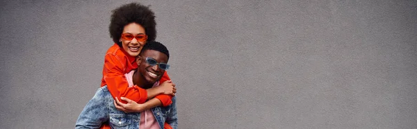 Positiva jovem afro-americana em óculos de sol e roupa brilhante piggybacking no melhor amigo e de pé perto do edifício na rua urbana, amigos da moda em ambientes urbanos, banner — Fotografia de Stock
