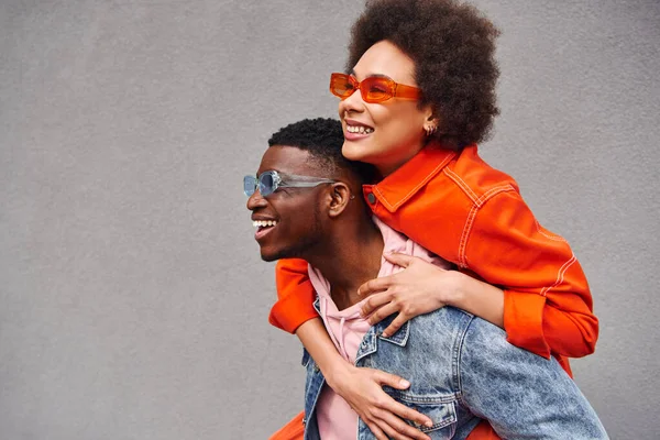 Junge und stylische afrikanisch-amerikanische Frau mit Sonnenbrille huckepack auf beste Freundin in Jeansjacke und Spaß in der Nähe des Gebäudes auf der urbanen Straße, trendige Freunde im urbanen Umfeld — Stockfoto