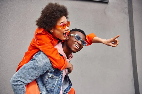 Aufgeregte junge afrikanisch-amerikanische Frau in Sonnenbrille und stylischem Outfit, die mit dem Finger zeigt, während sie huckepack auf Freund zeigt und in der Nähe von Gebäuden auf der städtischen Straße steht, trendige Freunde in städtischen Umgebungen — Stockfoto