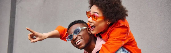 Jeune femme afro-américaine excitée avec des lunettes de soleil pointant du doigt tout en tirant sur le meilleur ami à la mode et debout près de la construction dans la rue urbaine, amis à la mode en milieu urbain, bannière — Photo de stock