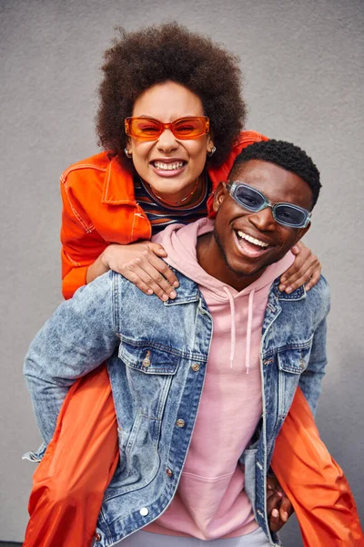 Portrait positiver und stilvoller junger afrikanisch-amerikanischer Frau mit Sonnenbrille huckepack auf besten Freund in Jeansjacke und Blick in die Kamera in der Nähe von Gebäuden im Freien, trendige Freunde in städtischen Umgebungen — Stockfoto