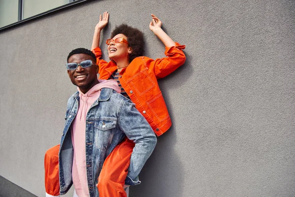 Happy young african american man in sunglasses and denim jacket holding best friend in bright outfit and having fun near wall on urban street, trendy friends in urban settings — Stock Photo
