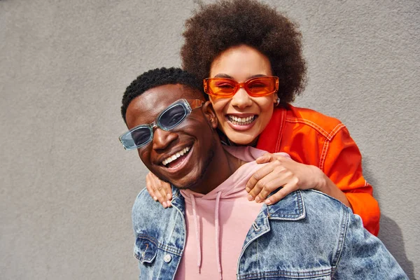 Портрет веселої молодої афроамериканки з натуральним волоссям, що обіймає найкращого друга в стильних сонцезахисних окулярах і джинсовій куртці, дивлячись на камеру біля будівлі, модні друзі в міських умовах — стокове фото