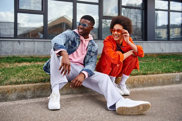 Full length of smiling and trendy young african american best friends in sunglasses looking away while sitting next to each other on border on blurred urban street, friends with trendy aesthetic — Stock Photo