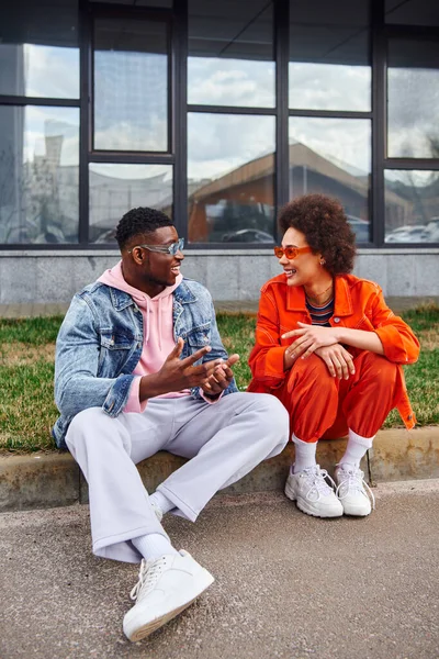 Jovens amigos afro-americanos alegres em óculos de sol e roupas brilhantes conversando enquanto passam o tempo e sentados na fronteira na rua urbana ao fundo, amigos com estética na moda — Fotografia de Stock