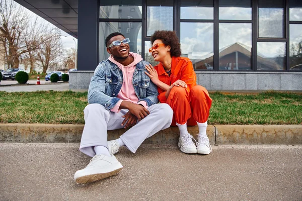 Une jeune femme afro-américaine positive portant des lunettes de soleil et une tenue lumineuse parlant à sa meilleure amie tout en passant du temps ensemble et assise à la frontière dans une rue urbaine, des amis à l'esthétique tendance — Photo de stock