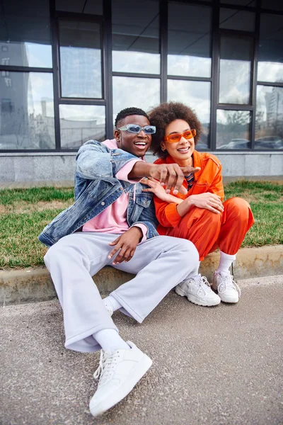 Smiling and trendy young african american man in sunglasses pointing with finger while sitting on border next to best friend in stylish outfit on urban street, friends with trendy aesthetic — Stock Photo