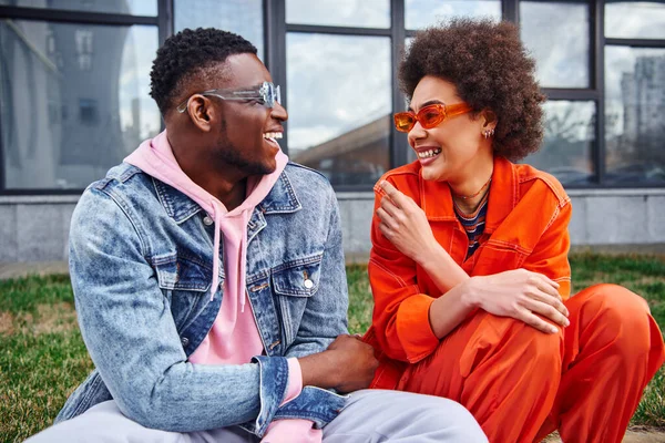 Jovens amigos afro-americanos alegres em óculos de sol e roupas elegantes conversando enquanto se sentam ao lado uns dos outros perto de um prédio borrado na rua urbana, amigos com estética na moda — Fotografia de Stock