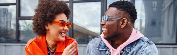 Lächelnde junge Afroamerikanerin in Sonnenbrille und hellem Outfit im Gespräch mit bester Freundin in Jeansjacke, während sie Zeit auf urbaner Straße verbringt, Freundinnen mit trendiger Ästhetik, Banner — Stockfoto