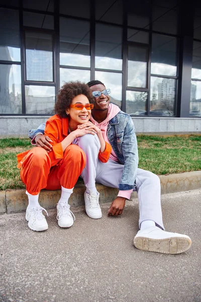 Comprimento total de positivo jovem afro-americano em óculos de sol abraçando melhor amigo em roupa brilhante e sentados juntos na fronteira na rua urbana, amigos com estética na moda — Fotografia de Stock