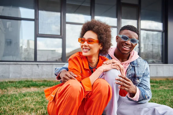 Fröhlicher junger und stilvoller afrikanisch-amerikanischer Mann mit Sonnenbrille umarmt beste Freundin in heller Kleidung und sitzt auf Gras in der Nähe verschwommener Gebäude auf der städtischen Straße, Freunde mit trendiger Ästhetik — Stockfoto