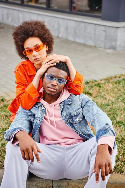 Модна молода афроамериканка в сонцезахисних окулярах і яскраве вбрання обіймає і позує з найкращим другом в джинсовій куртці і дивиться на камеру на міській вулиці, стильні друзі насолоджуються компанією — стокове фото