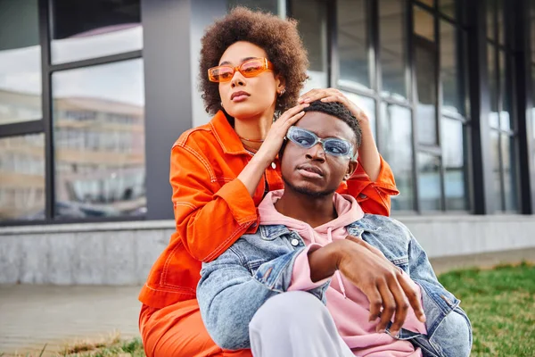 Confident and stylish young african american woman in sunglasses and bright outfit posing with best fried and looking away while spending time on urban street, stylish friends enjoying company — Stock Photo