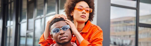 Confident and trendy young african american woman in sunglasses and bright outfit posing with best friend and looking away on blurred urban street, stylish friends enjoying company, banner — Stock Photo