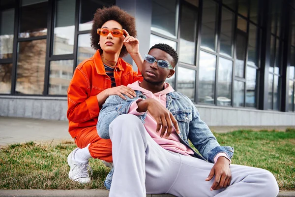 Stylish young african american woman in bright clothes wearing sunglasses and looking away near confident best friend sitting on border on urban street, stylish friends enjoying company — Stock Photo