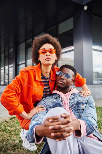 Trendy giovane donna afroamericana in occhiali da sole e vestito luminoso abbracciare elegante migliore amico e trascorrere del tempo su erba su strada urbana in background, amici alla moda godendo di compagnia — Foto stock