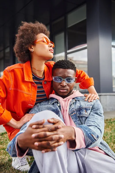 Confiant et moderne jeune femme afro-américaine étreignant meilleur ami à la mode dans les lunettes de soleil tout en passant du temps sur l'herbe dans la rue urbaine en arrière-plan, amis élégants profiter de la compagnie — Photo de stock