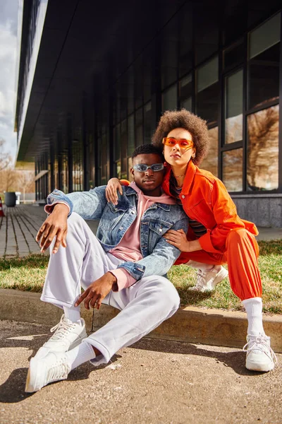 Full length of trendy and confident african american best friends in sunglasses looking at camera while sitting on border on blurred urban street at background, stylish friends enjoying company — Stock Photo