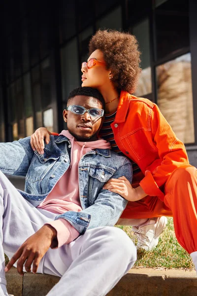 Modische junge afrikanisch-amerikanische Frau in Sonnenbrille und hellem Outfit umarmt beste Freundin sitzt an der Grenze auf verschwommenen städtischen Straße im Hintergrund, stilvolle Freunde genießen Gesellschaft — Stockfoto