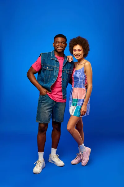 Full length of trendy and cheerful young african american best friends in summer clothes posing and looking at camera while standing on blue background, stylish friends radiating confidence — Stock Photo