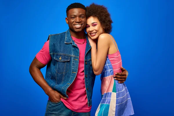 Cheerful and stylish young african american man in denim vest hugging best friend in sundress and looking at camera while standing isolated on blue, stylish friends radiating confidence — Stock Photo
