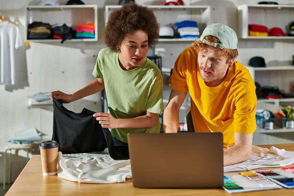 Young and multiethnic craftspeople looking at laptop while working together with clothes near coffee to go and color swatches on table in print studio, small business success concept — Stock Photo