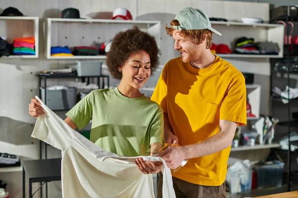 Smiling young craftsman holding printing layer near african american colleague with t-shirt while working together in blurred print studio at background, small business success concept — Stock Photo
