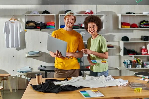 Positive young multiethnic craftspeople looking at camera while holding cloth swatches and laptop near clothes on table in print studio at background, small business success concept — Stock Photo