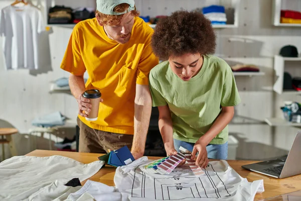 Joven diseñador afroamericano sosteniendo muestras de color cerca de la ropa mientras trabaja con un colega sosteniendo café para ir y portátil en la mesa en el estudio de impresión, próspero concepto de pequeña empresa - foto de stock