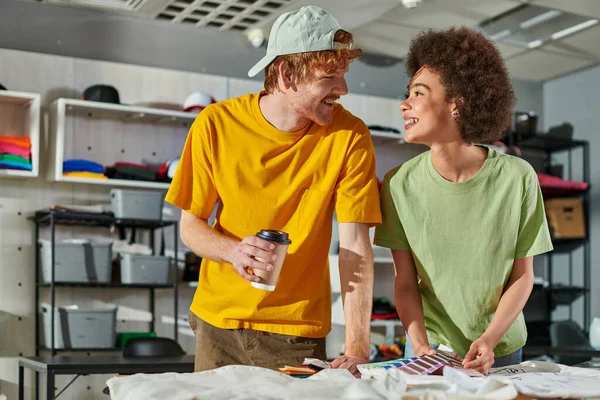 Cheerful multiethnic young designers talking while holding coffee to go and color swatches and working near clothes in blurred print studio at background, thriving small enterprise concept — Stock Photo