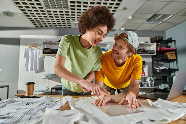 Smiling young african american craftswoman pointing with finger while working with colleague and printing layers near laptop in print studio, thriving small enterprise concept — Stock Photo