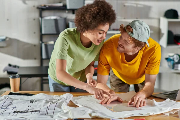 Positive young african american designer pointing with finger while working with colleague, printing layers and clothes near laptop in print studio, thriving small enterprise concept — Stock Photo