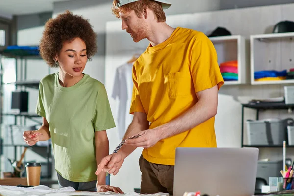 Young craftsman holding printing layer and talking to african american colleague while working together near clothes, coffee to go and laptop on table in print studio, start-up innovation concept — Stock Photo