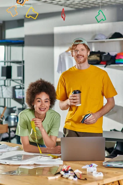Lächelnde junge multiethnische Designer mit Maßband und Coffee to go, während sie im Hintergrund in die Kamera neben Laptop und Kleidung auf dem Tisch im Druckstudio schauen, Innovationskonzept für Start-ups — Stockfoto