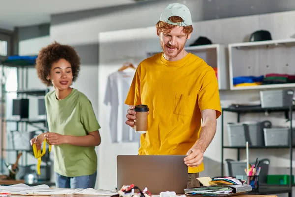 Smiling young designer holding coffee to go and using laptop while working near african american colleague with tape measure in blurred print studio, start-up innovation concept — Stock Photo