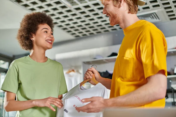Smiling young multiethnic designers talking and looking at each other while holding t-shirt with lettering on hanger and working in print studio, start-up innovation concept — Stock Photo
